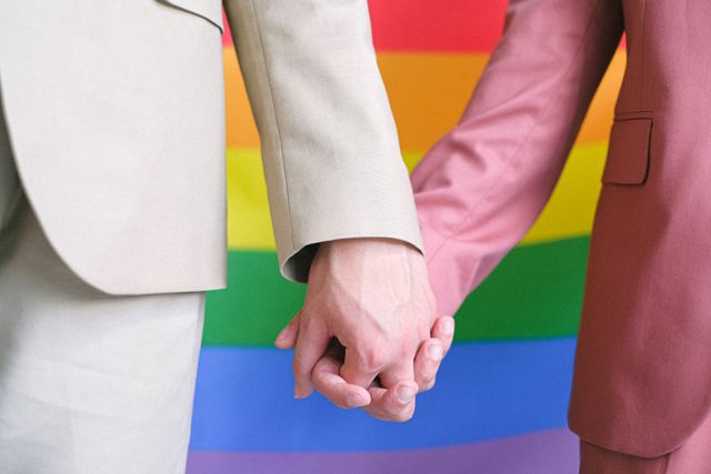 A close-up of a LGBTQ couple holding hands against a rainbow flag backdrop symbolizing unity and pride.